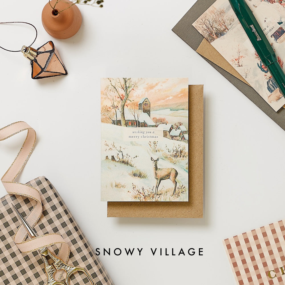 Christmas card with deer and snowy village design on a white surface with decorative items.