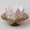 White paper crown with floral design on a gold stand against a white background