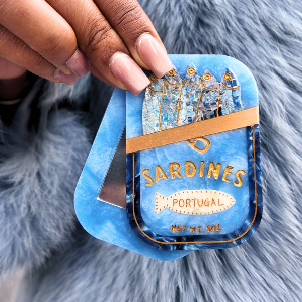 Blue decorative item resembling a sardine can with gold accents, held by a hand on a fluffy gray background.