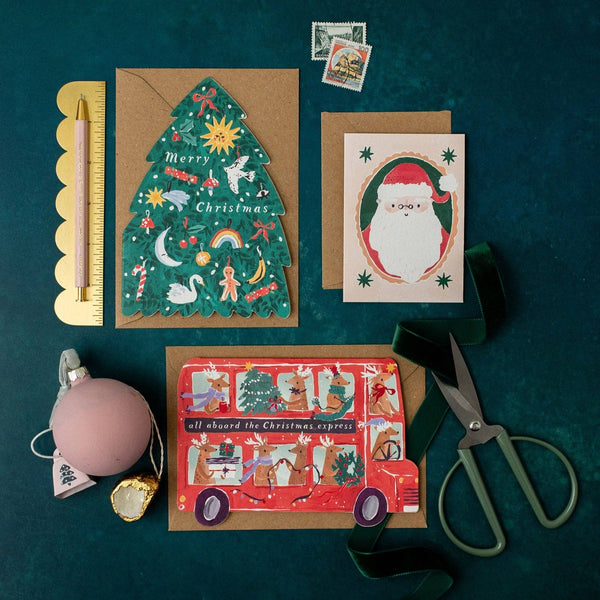Three Christmas-themed greeting cards on a dark green surface with stationery items.
