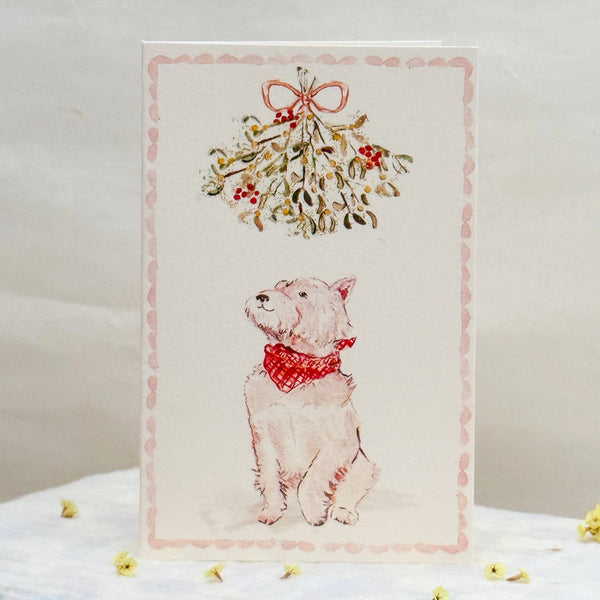 Card with a dog wearing a red bow and mistletoe design on a white surface.