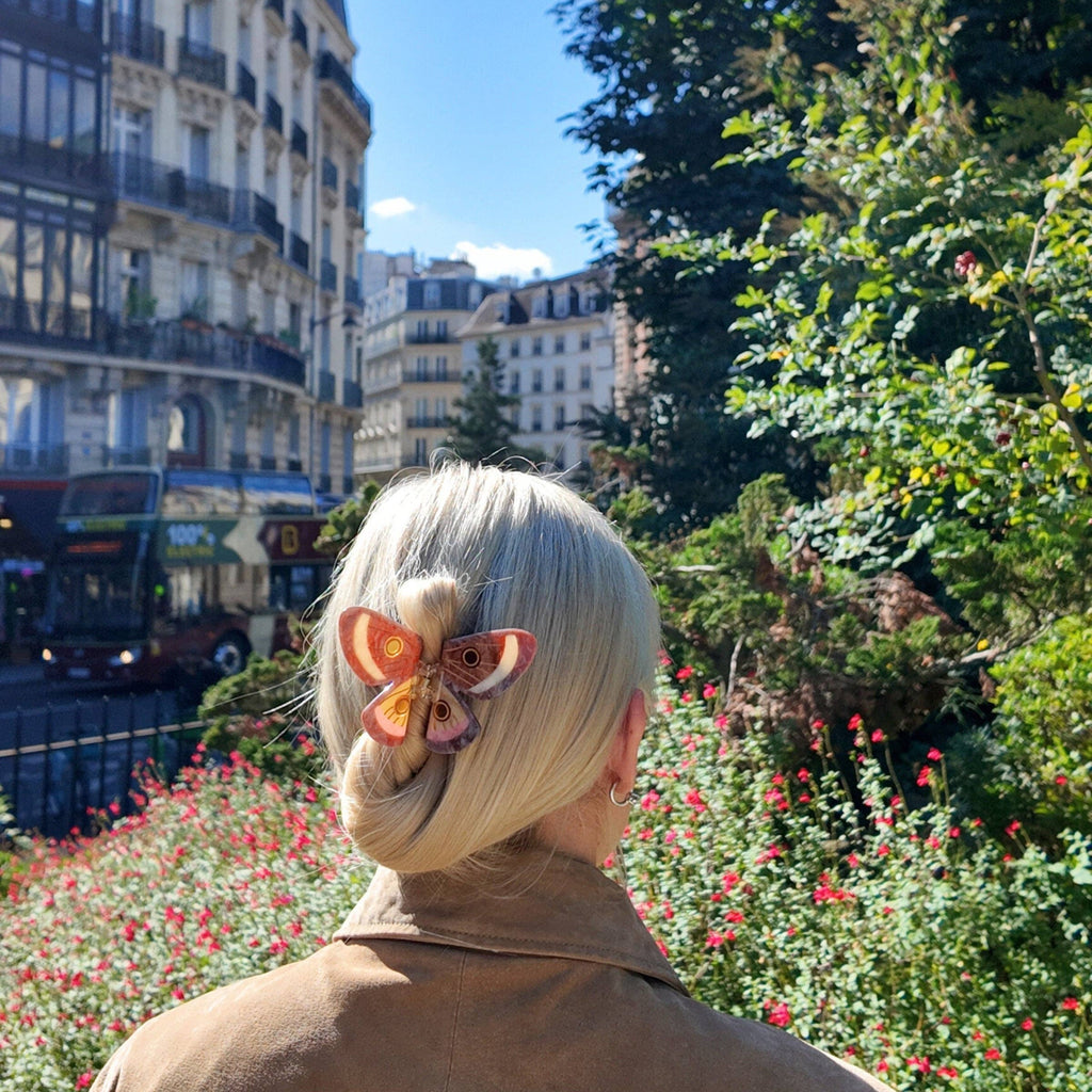 Person with a stylish haircut and butterfly hair accessory standing in a park with buildings and trees in the background.