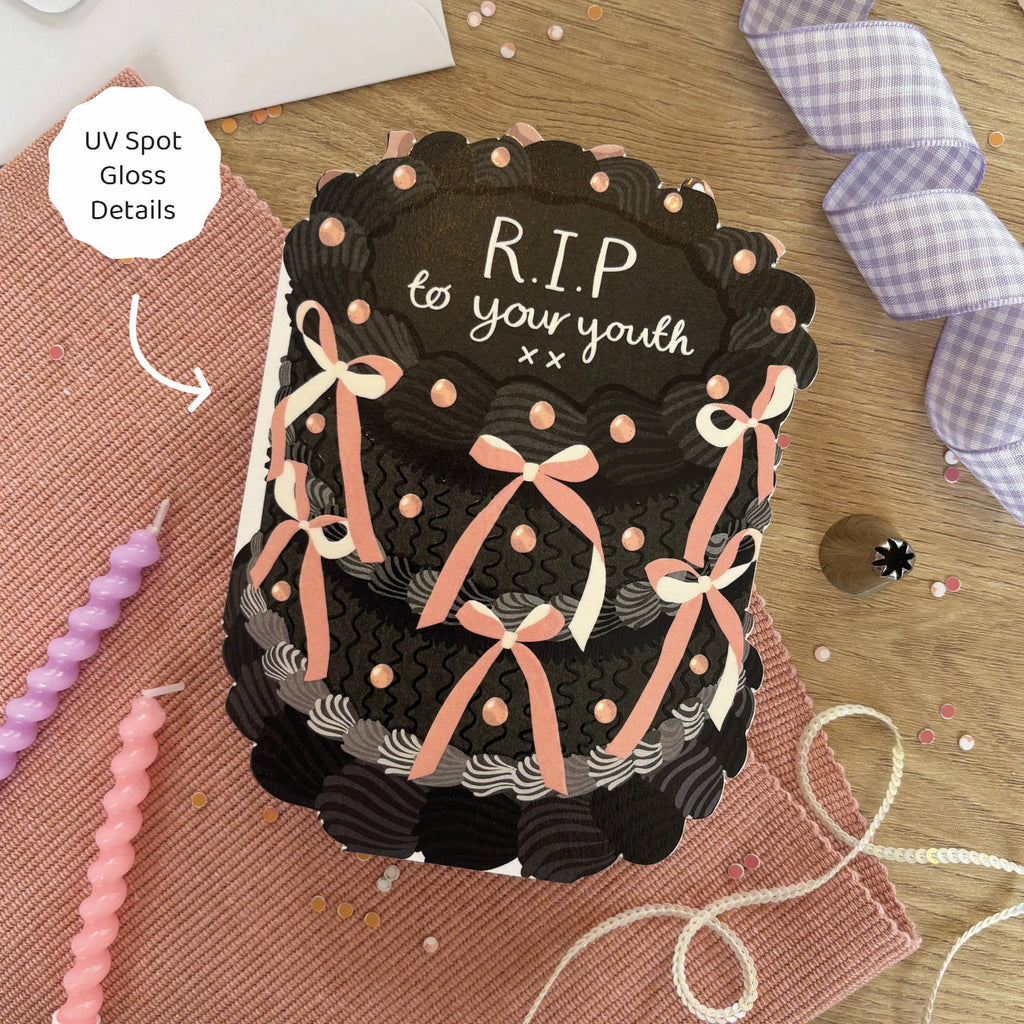 Black cake-shaped product with ribbons and 'R.I.P. to your youth' text on a wooden surface.