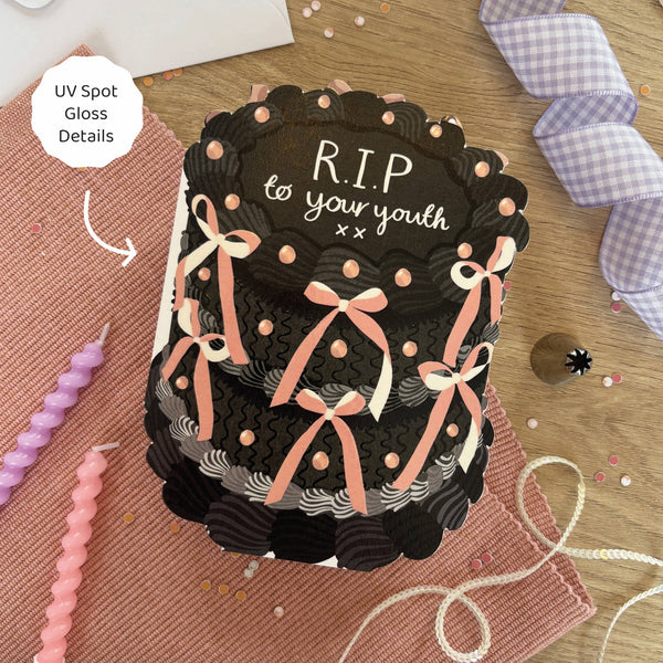 Black cake-shaped product with ribbons and 'R.I.P. to your youth' text on a wooden surface.