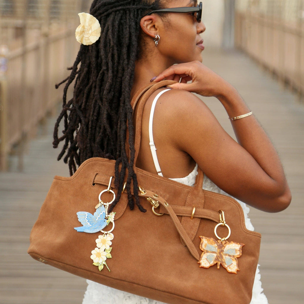 Woman with braided hair wearing a brown handbag with butterfly charms on a wooden bridge.