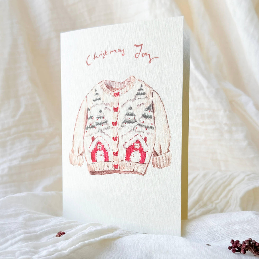 A vertical greeting card with a watercolor illustration of a Christmas sweater and the words 'Christmas Joy' at the top, standing upright on white textured fabric with small dried red berries nearby.