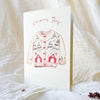 A vertical greeting card with a watercolor illustration of a Christmas sweater and the words 'Christmas Joy' at the top, standing upright on white textured fabric with small dried red berries nearby.