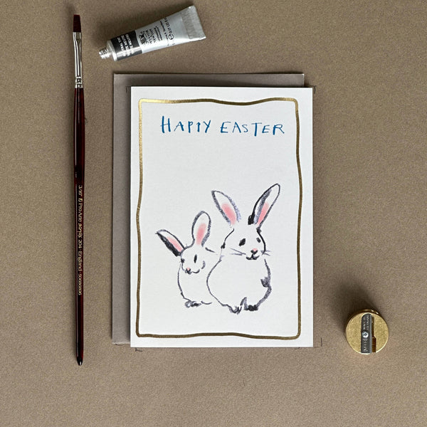 Greeting card with 'Happy Easter' text and rabbit illustrations, accompanied by a paintbrush and tube of paint on a brown background.