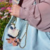 Person holding a light blue quilted handbag with a decorative charm featuring a penguin design, against a blurred floral background.