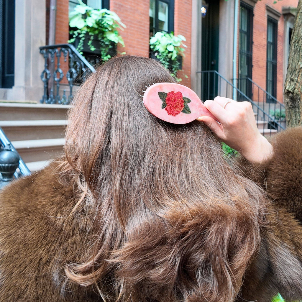 Person with wavy hair holding a pink hair clip with a rose design outdoors.