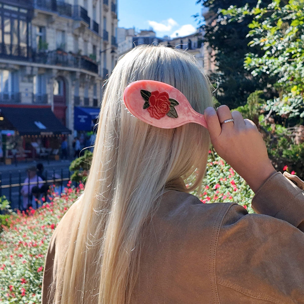 Person with a pink hairbrush with a rose design, standing outdoors with buildings and greenery in the background.