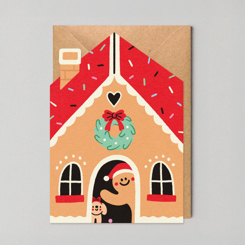 Gingerbread house card with a smiling face, wreath, and bow on a gray background