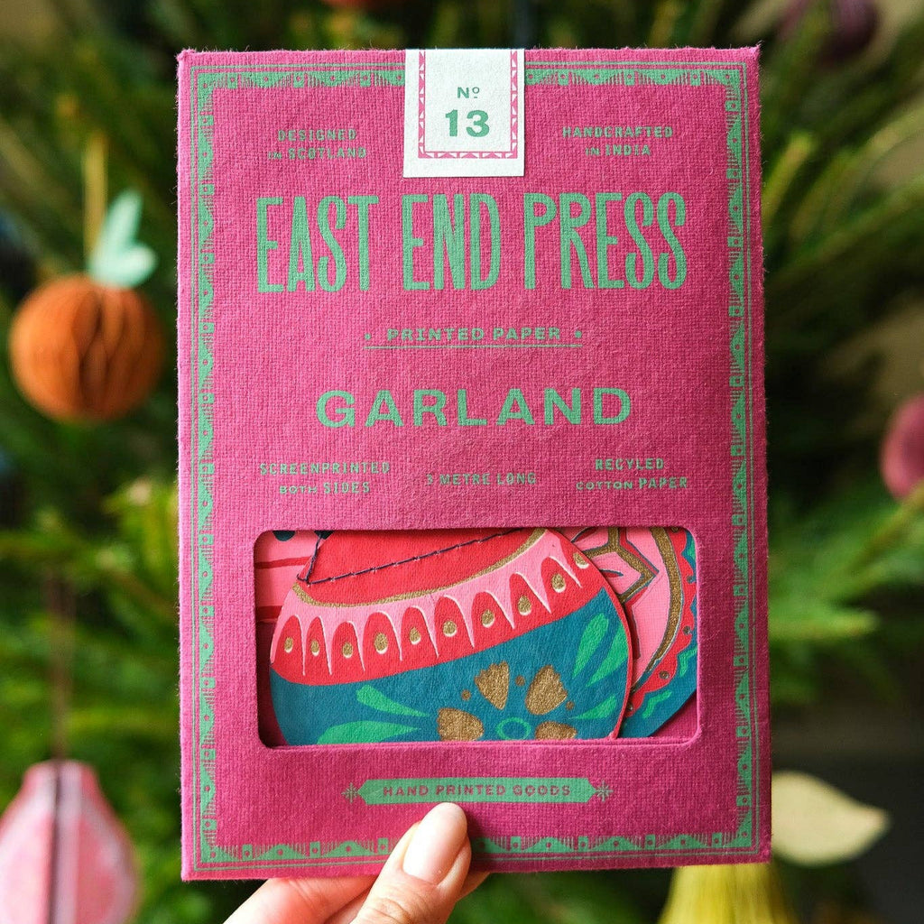 Pink packaging of East End Press Garland held in front of a decorative background with pumpkins and greenery.