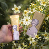Hand holding a tube of 'Shea Butter' with packaging amidst flowers