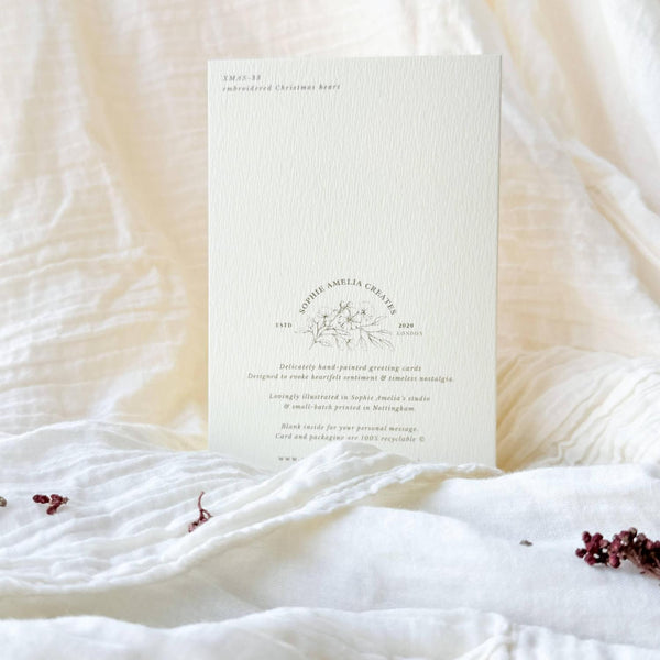 A vertical off-white invitation card with delicate gray text and floral line art, placed on soft white textured fabric with small dried flower petals scattered around it.