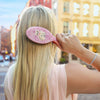Person with long blonde hair using a pink hairbrush in an urban setting