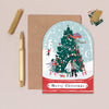 Christmas card with illustration of people decorating a tree, next to a pen and envelope on a beige background