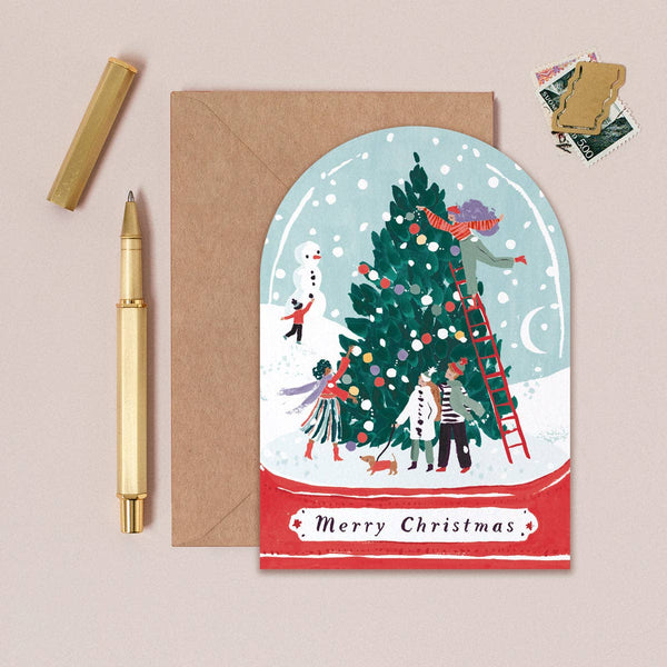 Christmas card with illustration of people decorating a tree, next to a pen and envelope on a beige background