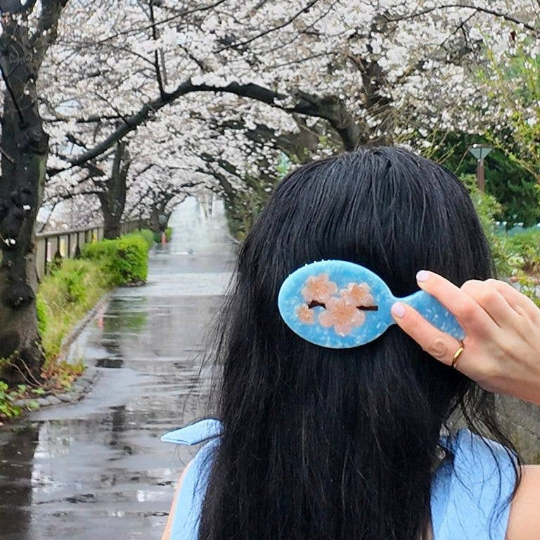 Person holding a blue hair clip with cherry blossoms in front of a cherry blossom tree-lined path.