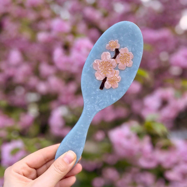 Blue hairbrush with cherry blossom design held in front of pink cherry blossoms