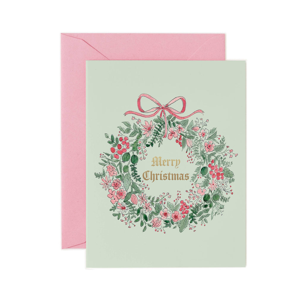 Christmas card with floral wreath and pink bow on a light green background, accompanied by a pink envelope.
