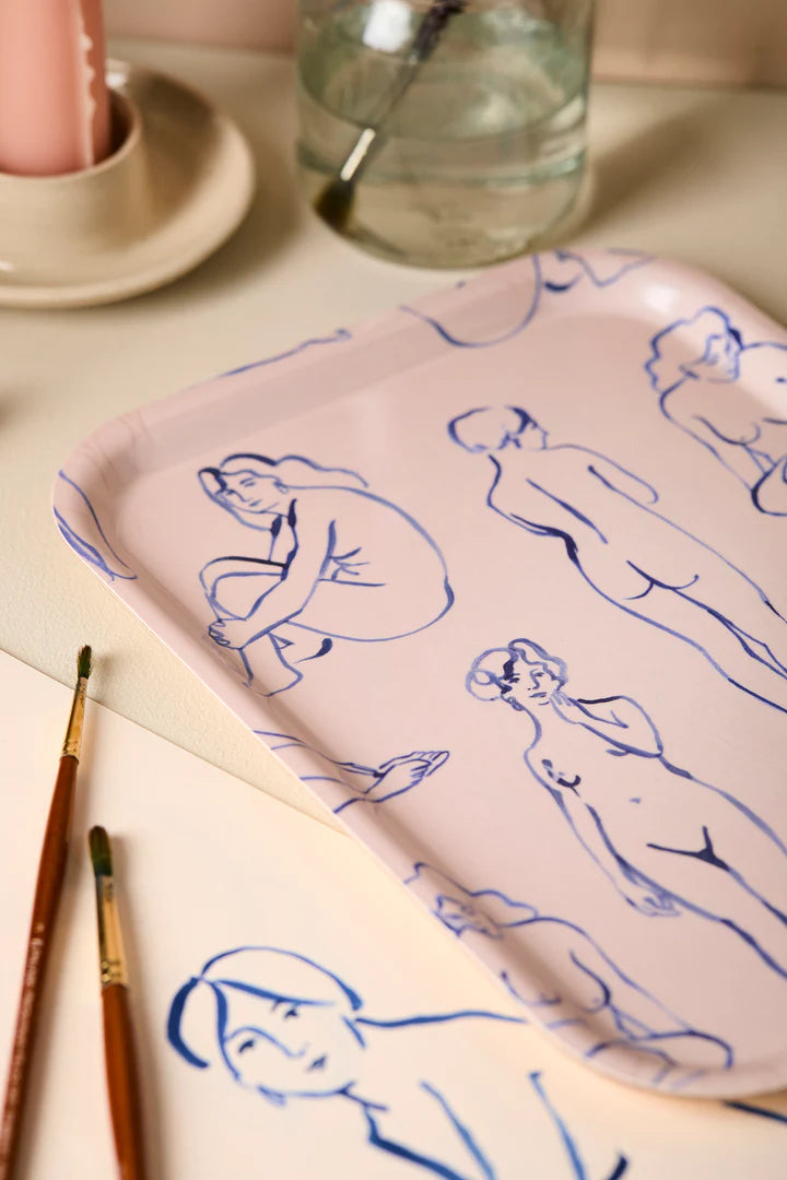 Pink tray with blue line drawings of women on a beige surface with brushes and water container.