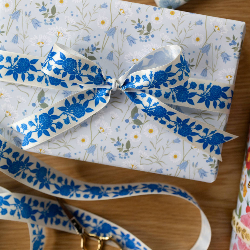 A gift box wrapped in white wrapping paper with a delicate blue and yellow floral print, tied with a matching blue floral ribbon, placed on a wooden surface.
