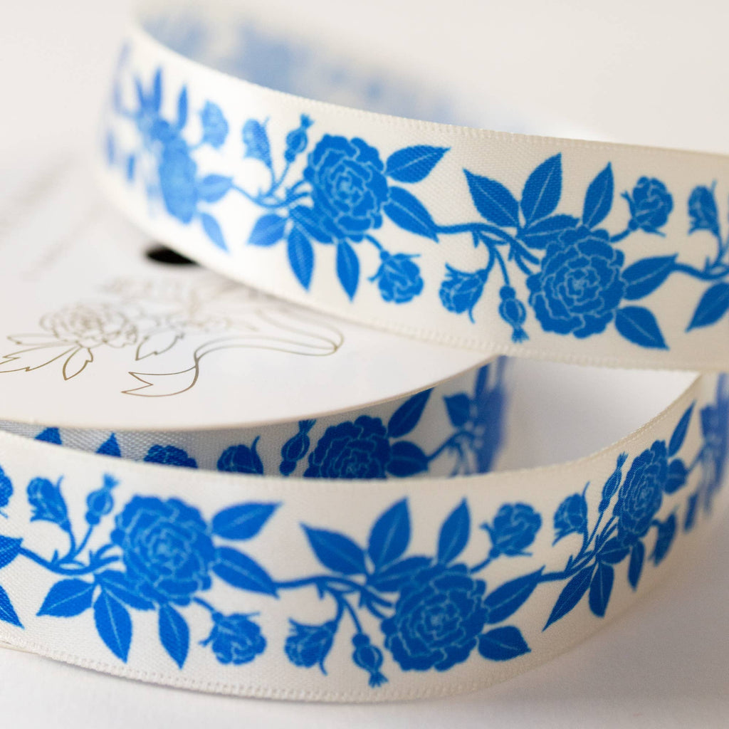 A close-up of a white ribbon with a repeating blue floral pattern, coiled on a light neutral background.