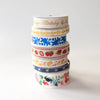 A stack of five decorative paper ribbons with various colorful patterns, including gold foil lettering, blue floral motifs, strawberries, yellow leaves, and multicolor botanical prints, set against a plain white background.