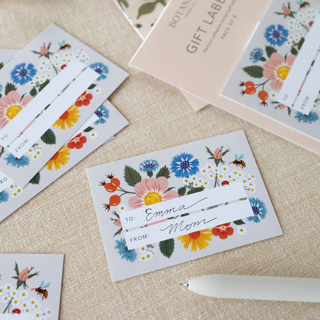 Botanica gift labels and matching gift tags with colorful floral illustrations, arranged on a beige fabric surface with a white pen nearby.