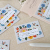 Botanica gift labels and matching gift tags with colorful floral illustrations, arranged on a beige fabric surface with a white pen nearby.