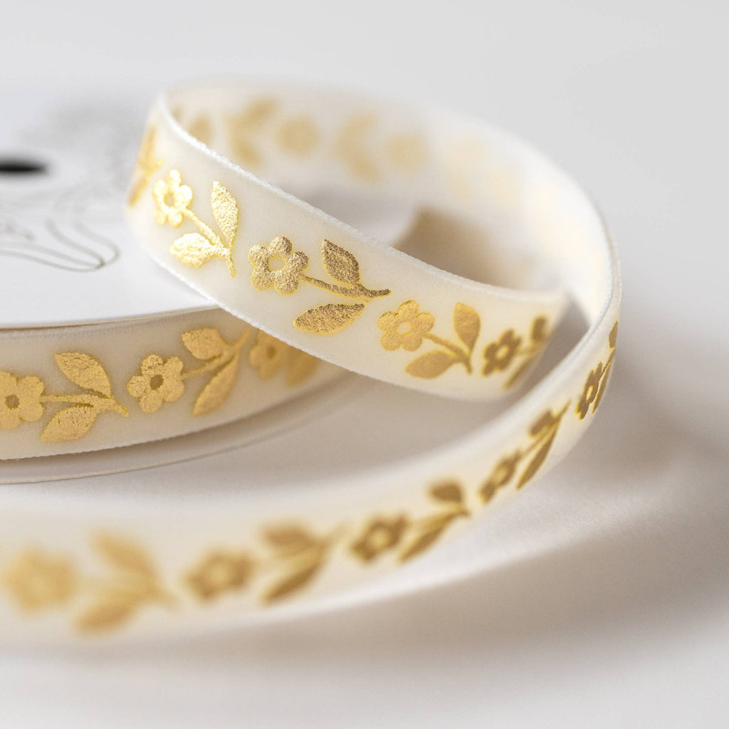 White ribbon with a delicate gold foil floral pattern, coiled on a white spool against a soft, light gray background.