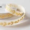 White ribbon with a delicate gold foil floral pattern, coiled on a white spool against a soft, light gray background.