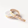 A roll of white washi tape with a gold foil floral pattern, partially unrolled from its white cardboard spool against a clean white background.
