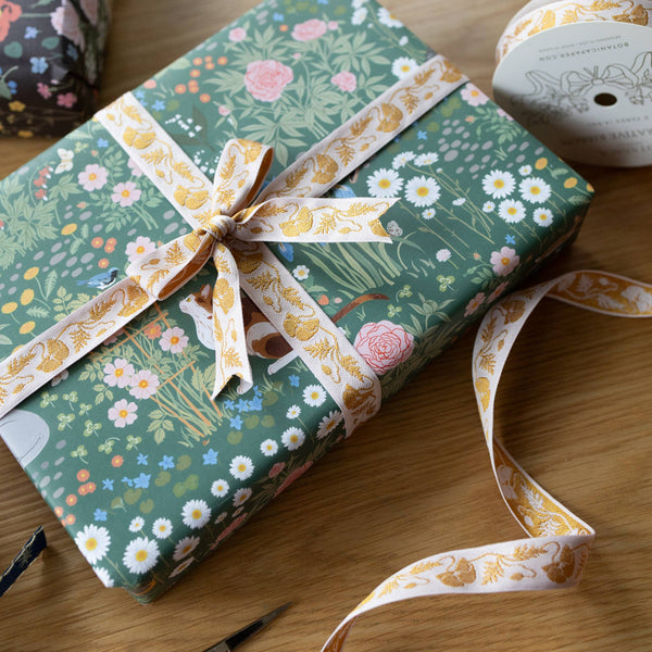 A wrapped gift box on a wooden table, covered in green floral wrapping paper with a cream ribbon printed with gold botanical motifs and tied in a bow. A spool of the same ribbon is visible in the upper right corner.