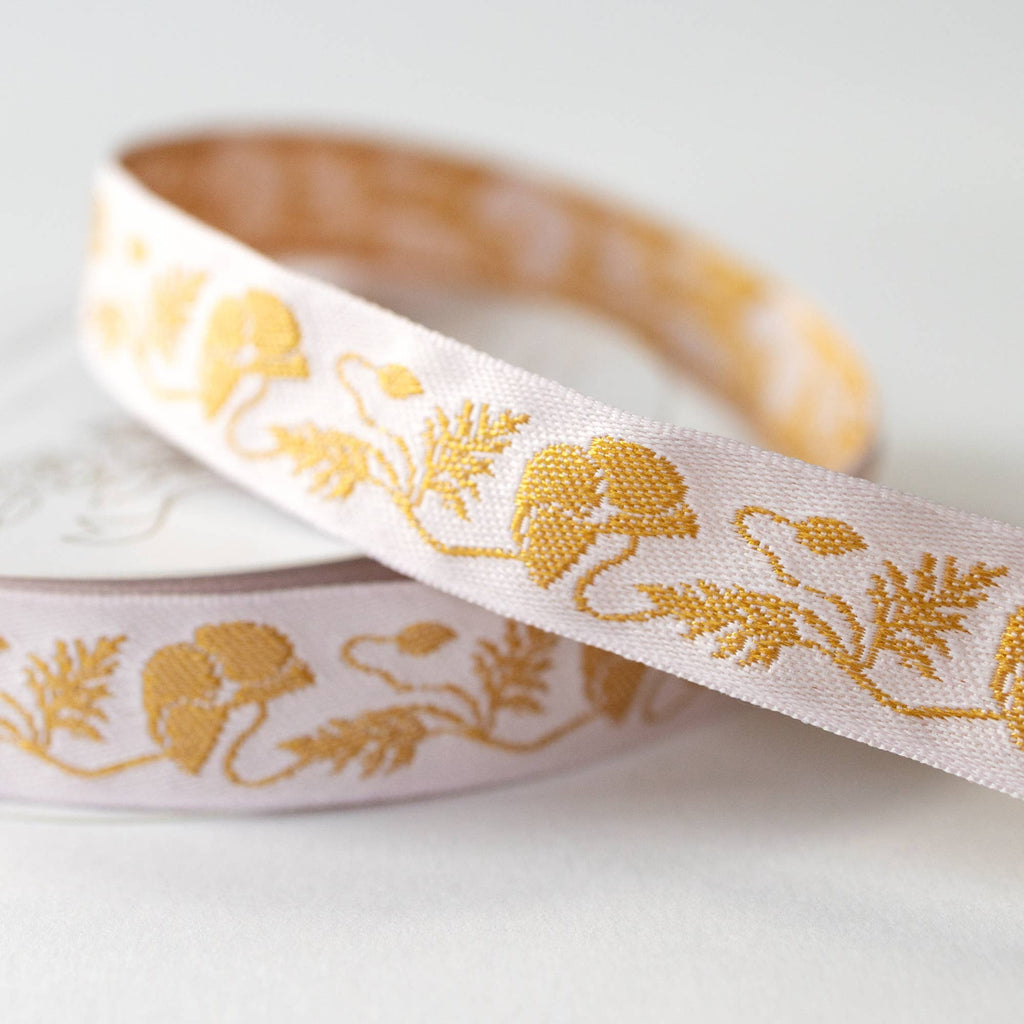 A close-up of a white ribbon with a repeating gold floral pattern, draped over a spool on a light gray background.