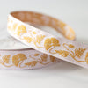 A close-up of a white ribbon with a repeating gold floral pattern, draped over a spool on a light gray background.