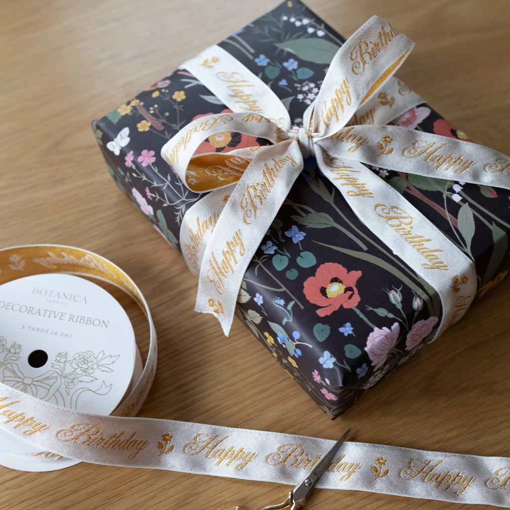 A gift box wrapped in floral botanical paper with a white satin ribbon printed with gold 'Happy Birthday' text, shown on a wooden surface with a spool of the ribbon and scissors nearby.