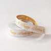 A spool of white satin ribbon with elegant gold foil 'Happy Birthday' script and small gold floral motifs, photographed on a clean white background.