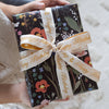 A wrapped gift box held by two hands, featuring a black floral patterned wrapping paper with orange, pink, and green botanical illustrations, tied with a cream ribbon printed with 'Happy Birthday' in gold script.