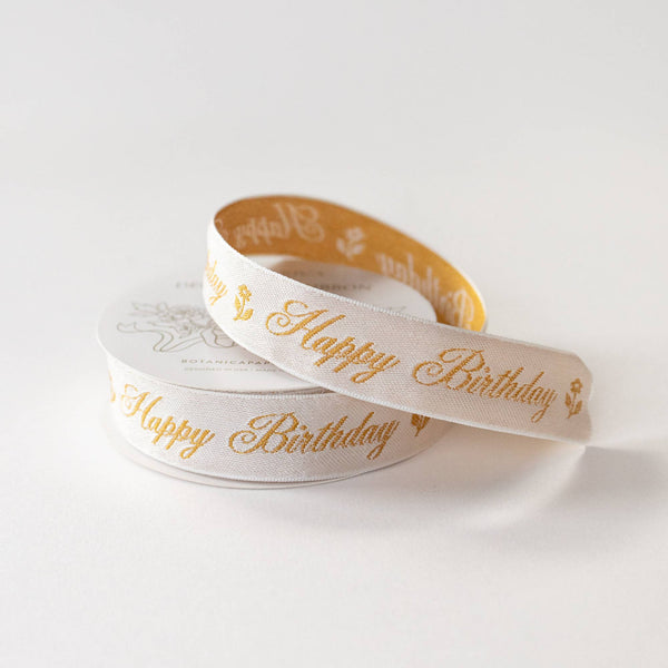 A spool of white satin ribbon with elegant gold foil 'Happy Birthday' script and small gold floral motifs, photographed on a clean white background.