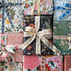 A flat-lay arrangement of multiple wrapped gift boxes featuring various colorful floral and botanical patterns, tied together with a cream satin ribbon printed with gold cursive text.