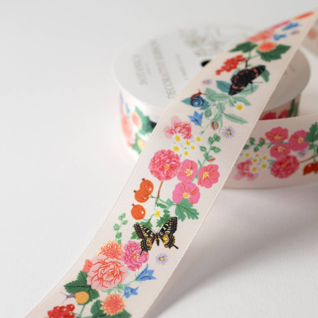 A close-up of a floral washi tape roll on a clean white background. The tape features a colorful pattern of pink flowers, green leaves, small birds, butterflies, and ladybugs on a cream-colored base.