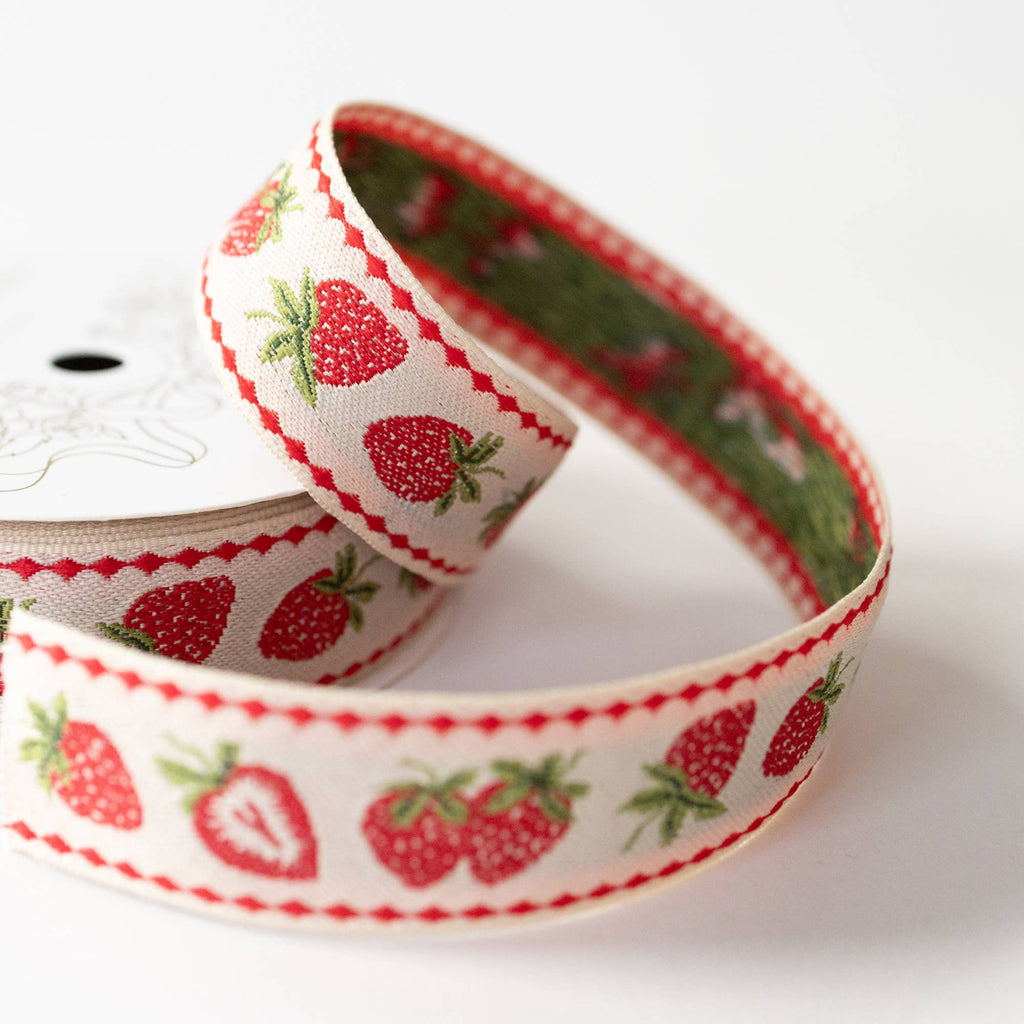 A close-up of a spool of grosgrain ribbon featuring a red and green strawberry pattern on a white background, with a red dotted border.