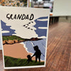 Card with illustration of a silhouette of a child and a baby, with 'GRANDAD' text above, on a wooden surface.