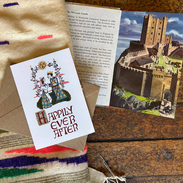 Card with 'Happily Ever After' text on a wooden surface with a castle postcard and fabric.