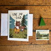 Greeting card with bears and 'Love you Dad' text on a wooden surface with a small postcard.