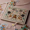 A square decorative sampler card or art print with a whimsical embroidered woodland scene, displayed on a dusty pink surface with teddy bear and star garlands nearby.