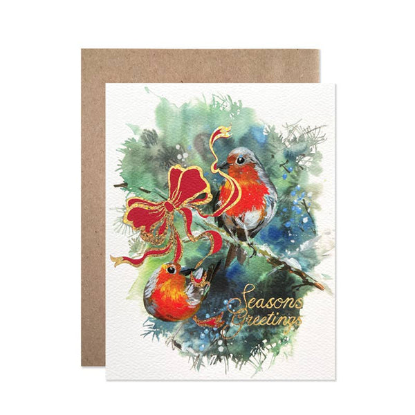 A square greeting card with a brown kraft envelope behind it. The card features a watercolor illustration of two red-breasted birds among green and blue foliage, with a large red bow and gold cursive text reading 'Seasons Greetings'.