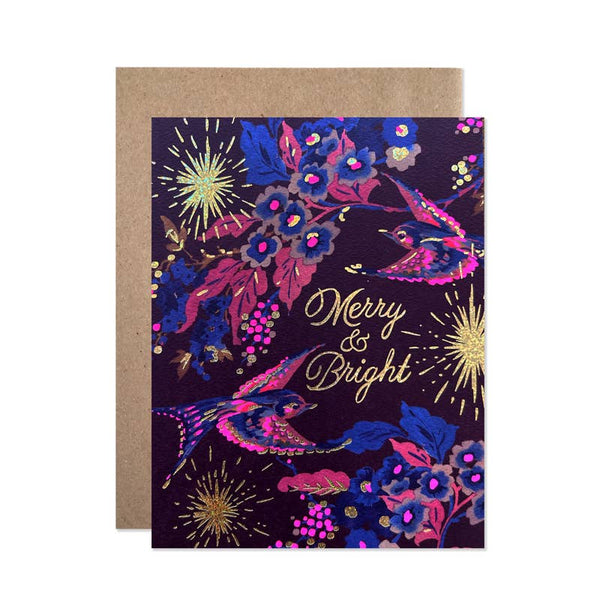 A festive greeting card with a dark purple background, decorated with pink and blue birds, gold fireworks, and the words 'Merry & Bright' in gold script, shown with a brown kraft envelope behind it.